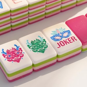 May include: Close-up of colorful Mahjong tiles. The tiles have a white top with various designs, including a pink dragon, a green face, and a blue mask with the word "JOKER". The sides are striped with pink and green. The tiles are arranged in rows.