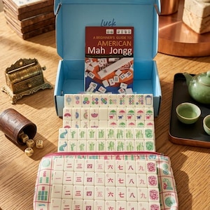 May include: A Mah Jongg game set featuring a light blue storage box, a beginner's guide booklet, and numerous white tiles with colorful symbols, numbers, and characters. The set includes a patterned fabric pouch and a wooden dice cup with dice.