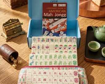 May include: A Mah Jongg game set featuring a light blue storage box, a beginner's guide booklet, and numerous white tiles with colorful symbols, numbers, and characters. The set includes a patterned fabric pouch and a wooden dice cup with dice.