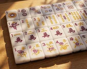 May include: A close-up of a Mahjong set with white tiles. The tiles feature various symbols and numbers in red and gold. The set is arranged on a wooden surface, with some tiles displaying floral designs.