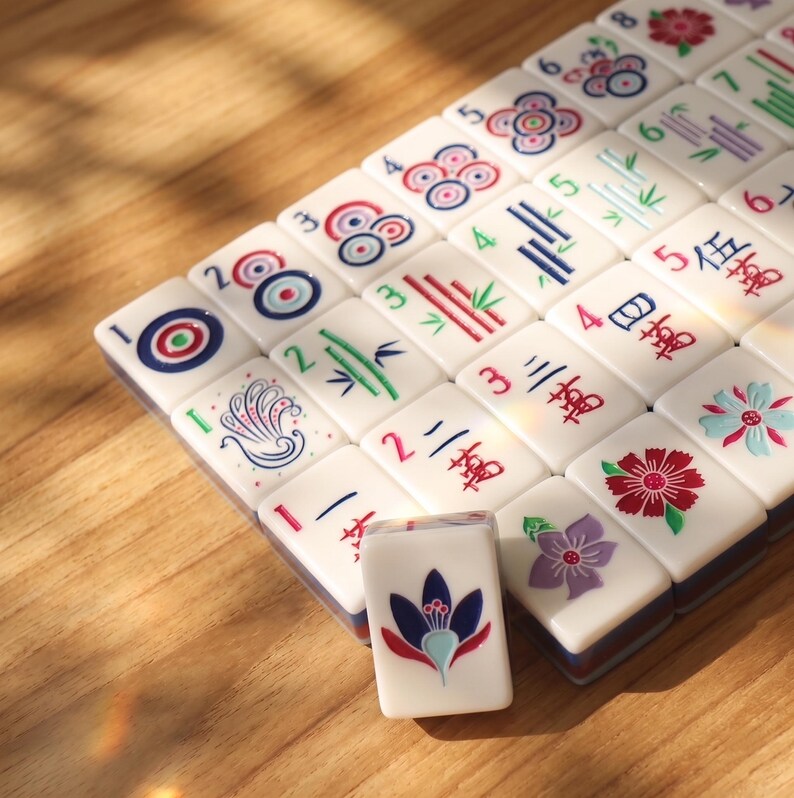 May include: A set of white Mahjong tiles with various colorful symbols and numbers. The tiles feature red, blue, green, and purple designs, including flowers, bamboo, and circles. One tile is standing upright, showcasing a blue and red floral design.