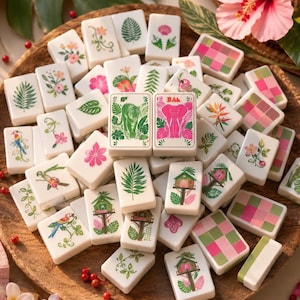 Op de afbeelding: Een verzameling witte rechthoekige mahjongtegels met kleurrijke illustraties van tropische planten, bloemen, vogels, boomhutten en geometrische patronen. Twee centrale tegels tonen een groene tijger en een roze olifant.