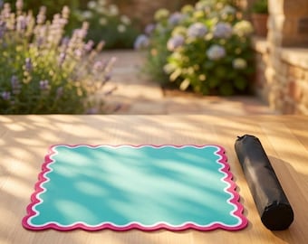 May include: A rectangular placemat with a solid turquoise center, a white border, and a scalloped pink edge, accompanied by a black cylindrical carrying case. The placemat features a smooth surface and is positioned flat on a wooden surface.