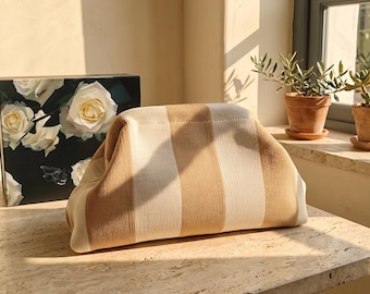 May include: A soft-sided clutch bag featuring a vertical striped pattern in alternating shades of beige and cream. The bag has a rounded, structured top opening and a textured knit fabric construction, presented in a neutral color palette.