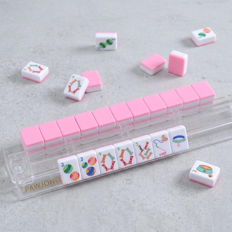 May include: A pink and white PawJong set with tiles featuring colourful designs and the word "EAST". The tiles are arranged in a clear plastic tray. Some tiles are scattered around the tray.