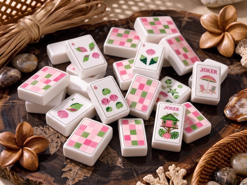 May include: A collection of white Mahjong tiles featuring pink and green checkered backs and various tropical-themed illustrations on the fronts, including seashells, a beach umbrella with chairs, and a small house, all arranged in a scattered pile.