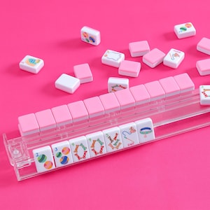 May include: A Mahjong set with pink and white tiles, and a clear plastic tile holder. The tiles have colourful illustrations. The background is a solid pink.