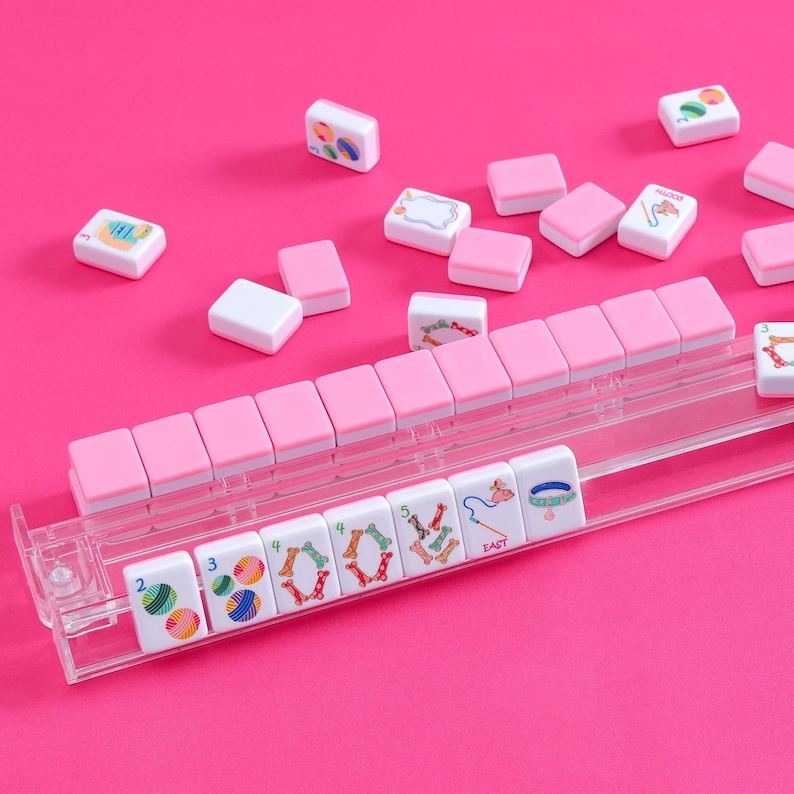 May include: A Mahjong set with pink and white tiles, and a clear plastic tile holder. The tiles have colourful illustrations. The background is a solid pink.