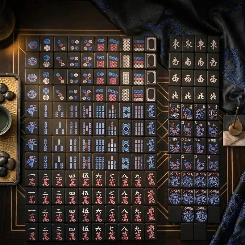 May include: A set of black Mahjong tiles featuring engraved characters, numbers, and symbols in red, blue, and white. The tiles include suits of dots, bamboo, and characters, along with wind tiles, dragon tiles, flower tiles, and Joker tiles.