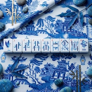 Puede incluir: Azulejos de Mahjong blancos con ilustraciones azules, incluyendo bamb&uacute;, una grulla y otros s&iacute;mbolos. Los azulejos est&aacute;n dispuestos sobre una tela con estampado Chinoiserie azul y blanco, con acentos de bamb&uacute; y piedras decorativas.