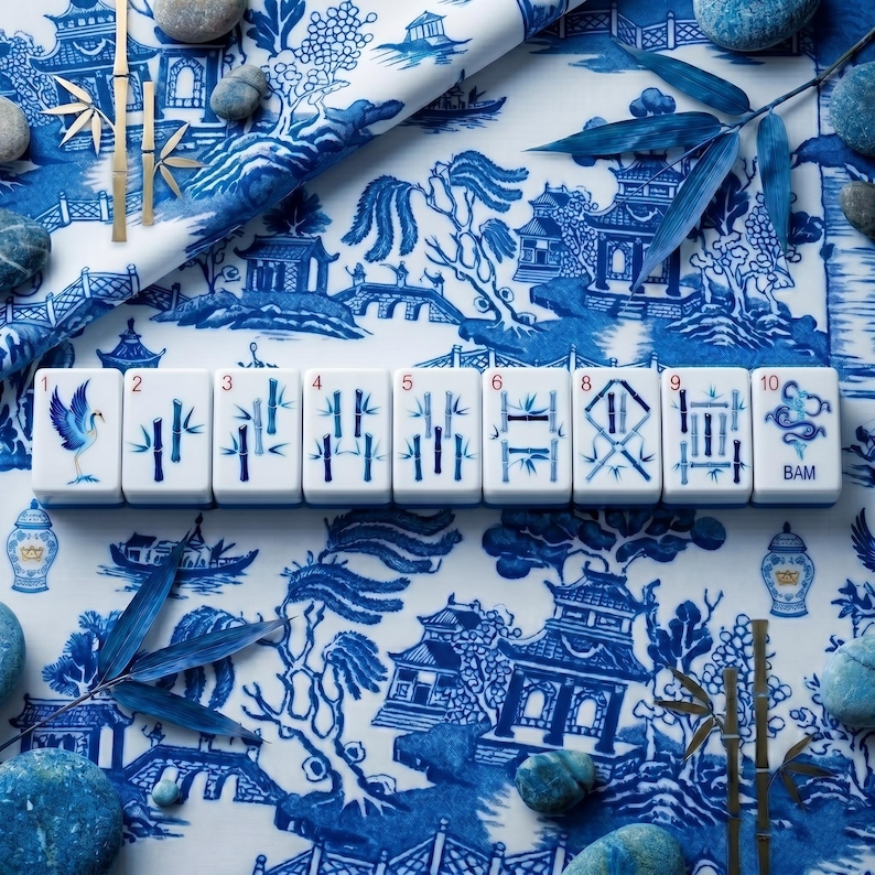 Puede incluir: Azulejos de Mahjong blancos con ilustraciones azules, incluyendo bamb&uacute;, una grulla y otros s&iacute;mbolos. Los azulejos est&aacute;n dispuestos sobre una tela con estampado Chinoiserie azul y blanco, con acentos de bamb&uacute; y piedras decorativas.
