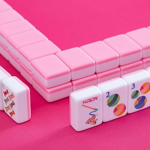 May include: A Mahjong set in pink and white, displayed on a pink surface. The tiles are white with pink edges, displaying colourful illustrations and numbers. The set is composed of multiple tiles, creating a visually appealing arrangement.