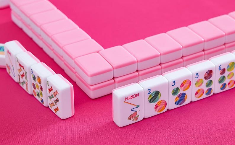 May include: A Mahjong set in pink and white, displayed on a pink surface. The tiles are white with pink edges, displaying colourful illustrations and numbers. The set is composed of multiple tiles, creating a visually appealing arrangement.