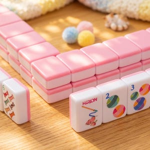 May include: Pink and white Mahjong tiles arranged on a wooden surface. The tiles have pink edges and white faces with colorful illustrations and numbers. The word "HION" is visible on one tile. Small, colorful pom-poms are scattered around the tiles.