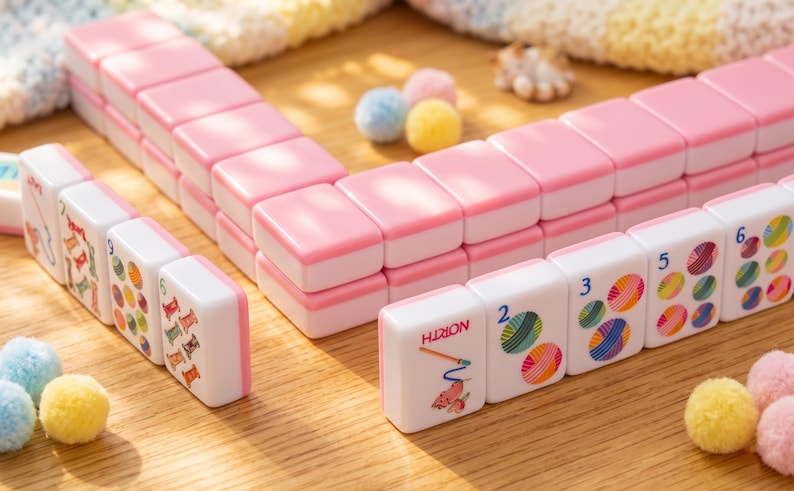 May include: Pink and white Mahjong tiles arranged on a wooden surface. The tiles have pink edges and white faces with colorful illustrations and numbers. The word "HION" is visible on one tile. Small, colorful pom-poms are scattered around the tiles.
