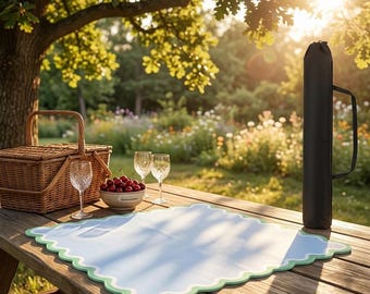 May include: A light blue square placemat with a scalloped light green border, a woven wicker picnic basket, two clear glass goblets, a small bowl filled with red cherries, and a tall black cylindrical carrying case with a handle.