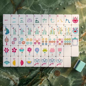 Może przedstawiać: Kompletny zestaw białych płytek do gry w Mahjong z r&oacute;żnymi symbolami i chińskimi znakami. Płytki przedstawiają smoki, kwiaty, bambus i liczby. Mały stos płytek i moneta są widoczne na zielonej marmurowej powierzchni.
