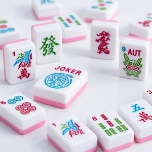 May include: A collection of white Mahjong tiles with pink bases, featuring various colorful engraved symbols, Chinese characters, and the word JOKER in red text. The tiles display numbers, dragon motifs, and floral patterns in blue, green, and red ink.