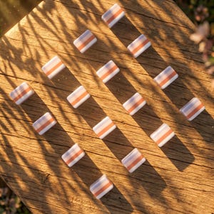 May include: An overhead shot of rectangular, white objects with orange and light purple stripes. The objects are arranged in a grid pattern on a wooden surface. The scene is lit with natural sunlight, casting shadows.