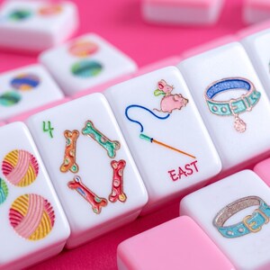 May include: Close-up of white Mahjong tiles with colourful illustrations and the word "EAST" in red. The tiles feature images of a mouse, bones, and a cat collar. The background is a vibrant pink.