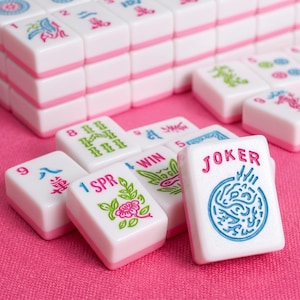 May include: A set of white Mahjong tiles with pink bases, featuring colorful engraved designs including numbers, Chinese characters, floral patterns, and the text JOKER, WIN, and SPR, arranged in a stack and scattered on a textured pink surface.
