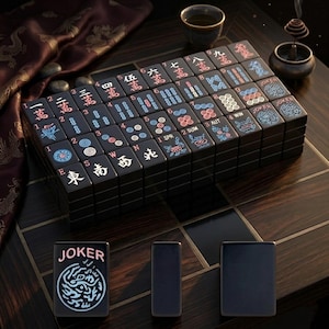 May include: A set of black mahjong tiles featuring engraved characters, numbers, and symbols in red, blue, and white. The tiles are arranged in a rectangular block, with three individual tiles placed in front, including one labeled JOKER.