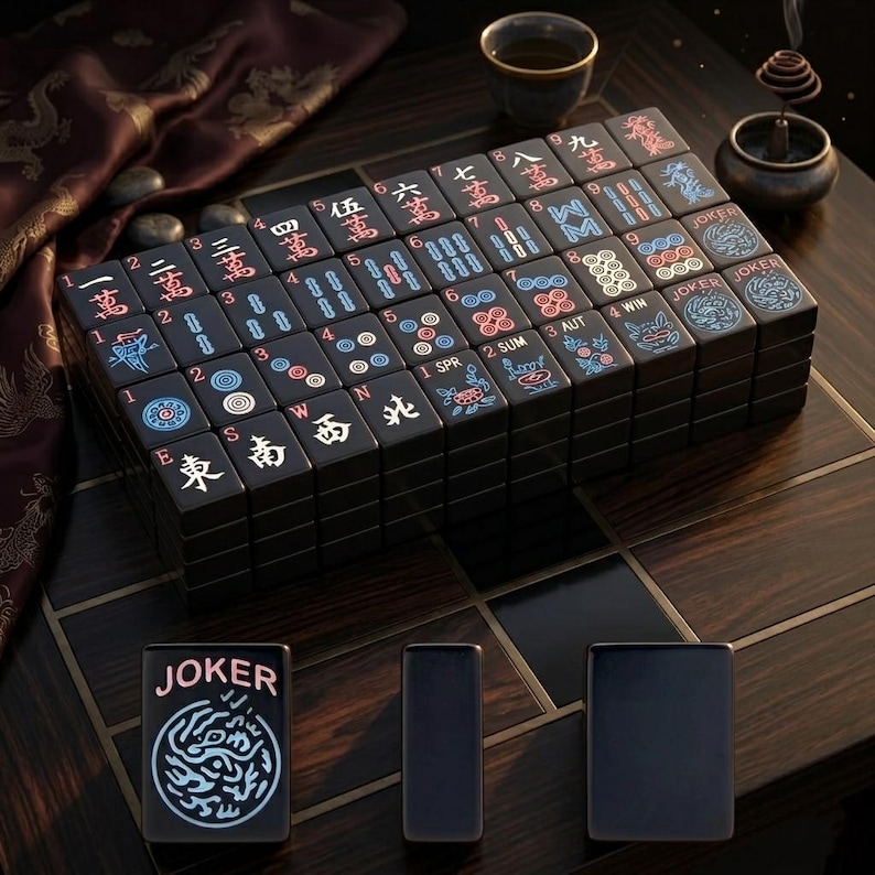 May include: A set of black mahjong tiles featuring engraved characters, numbers, and symbols in red, blue, and white. The tiles are arranged in a rectangular block, with three individual tiles placed in front, including one labeled JOKER.