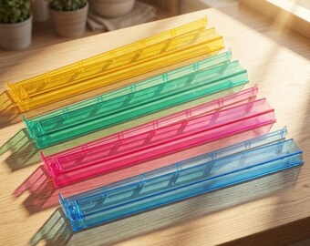May include: A set of four long, narrow, rectangular storage trays made of translucent plastic in yellow, green, pink, and blue. Each tray features a flat base with raised side walls and small vertical dividers along the interior length.