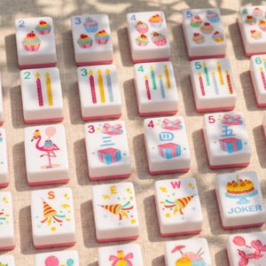 May include: A collection of white Mahjong tiles with pink edges, featuring colorful illustrations. Designs include cupcakes, candles, gift boxes, a flamingo, and party horns. Some tiles display numbers, letters, and the word "JOKER".