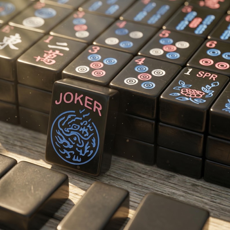 May include: A set of black Mahjong tiles with engraved designs in blue and pink. One tile stands upright in the foreground, featuring the word JOKER in pink above a blue circular dragon graphic. Other tiles display numbers, floral patterns, and symbols.