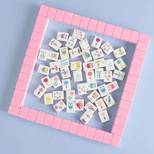 May include: A collection of white mahjong tiles with colorful illustrations, including flowers, anchors, and luggage, are scattered within a pink rectangular frame. The tiles are arranged on a light blue surface, creating a visually appealing game set.
