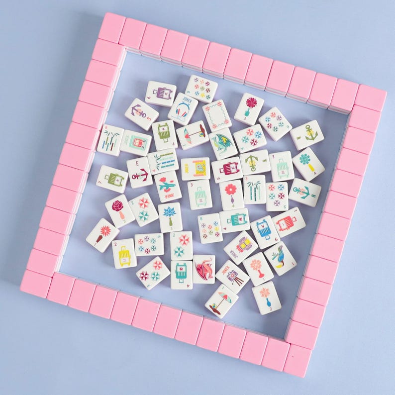 May include: A collection of white mahjong tiles with colorful illustrations, including flowers, anchors, and luggage, are scattered within a pink rectangular frame. The tiles are arranged on a light blue surface, creating a visually appealing game set.
