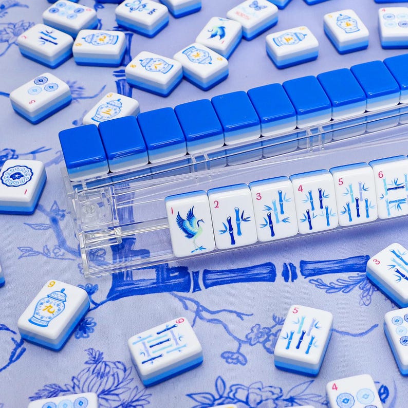 Puede incluir: Un conjunto de fichas de Mahjong blancas y azules, con varios s&iacute;mbolos y n&uacute;meros. Las fichas est&aacute;n dispuestas sobre una superficie con estampado floral azul. Se ve un soporte de fichas de pl&aacute;stico transparente. Las fichas tienen una combinaci&oacute;n de colores azul y blanco.
