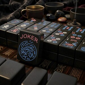 May include: A set of black mahjong tiles with engraved designs in blue, red, and white. One tile stands upright in the foreground, featuring the word JOKER in pink text above a blue circular dragon emblem. Other tiles display numbers, symbols, and floral patterns.