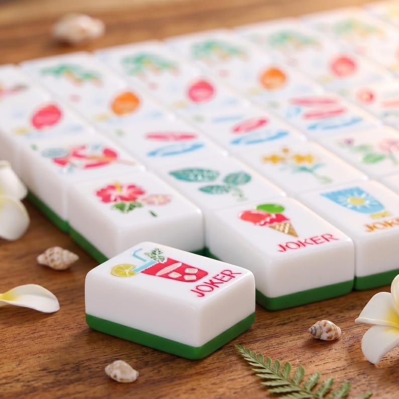 K&ouml;nnte beinhalten: Ein Satz wei&szlig;er Mahjong-Steine mit gr&uuml;nen Kanten, die farbenfrohe Illustrationen von Blumen, Fr&uuml;chten und Getr&auml;nken zeigen. Ein Stein zeigt einen Cocktail mit dem Wort "JOKER". Die Steine sind auf einer Holzoberfl&auml;che mit dekorativen Elementen angeordnet.