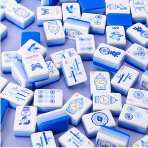 Puede incluir: Un conjunto de fichas de Mahjong blancas y azules. Las fichas presentan varios dise&ntilde;os, incluyendo tallos de bamb&uacute;, patrones florales y la palabra "NORTH". Las fichas est&aacute;n dispuestas en un patr&oacute;n disperso, con algunas fichas que muestran n&uacute;meros y s&iacute;mbolos.