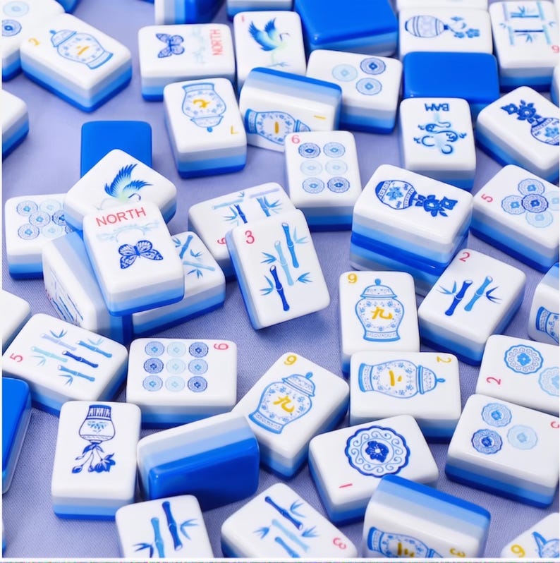 Puede incluir: Un conjunto de fichas de Mahjong blancas y azules. Las fichas presentan varios dise&ntilde;os, incluyendo tallos de bamb&uacute;, patrones florales y la palabra "NORTH". Las fichas est&aacute;n dispuestas en un patr&oacute;n disperso, con algunas fichas que muestran n&uacute;meros y s&iacute;mbolos.