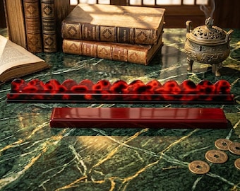 May include: A long, rectangular display stand with a scalloped top edge featuring a red and black tortoiseshell pattern, positioned in front of a solid, dark red rectangular base on a green marble surface with books and a metal incense burner.