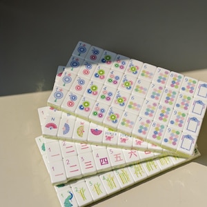 May include: A set of white rectangular Mahjong tiles arranged in three overlapping rows. The tiles feature colorful printed designs including numbers 1 through 9, directional symbols, bamboo stalks, peacock illustrations, and the text JOKER.