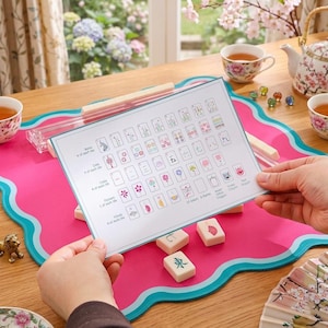 May include: A person holding a game guide over a pink and teal game mat. The guide displays various game tiles and symbols. Several ivory-colored game tiles are scattered on the mat. A tea set and decorative items are in the background.