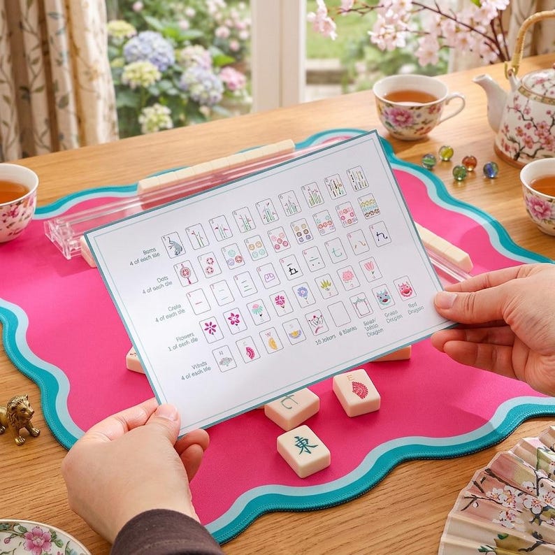May include: A person holding a game guide over a pink and teal game mat. The guide displays various game tiles and symbols. Several ivory-colored game tiles are scattered on the mat. A tea set and decorative items are in the background.