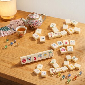 May include: A collection of ivory-colored Mahjong tiles with colorful illustrations and numbers, arranged on a wooden surface. A floral teapot, teacup, and decorative fan are also present, along with glass marbles. The scene suggests a game setup.