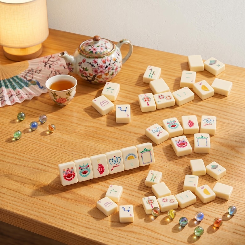 May include: A collection of ivory-colored Mahjong tiles with colorful illustrations and numbers, arranged on a wooden surface. A floral teapot, teacup, and decorative fan are also present, along with glass marbles. The scene suggests a game setup.