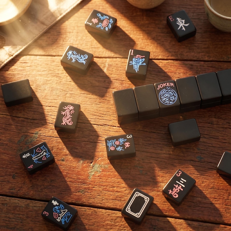 May include: A set of black rectangular mahjong tiles scattered on a wooden surface. The tiles feature engraved designs in blue, red, and white, including Chinese characters, floral patterns, dragon motifs, and one tile clearly labeled JOKER.