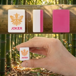 May include: A white and pink Mahjong tile with a bee design and the word "JOKER". The tile measures 0.9" x 1.2" x 0.5". The bee is orange with a crown. The background is bamboo.