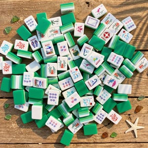 Op de afbeelding: Een verzameling groene en witte Mahjong-tegels verspreid over een houten oppervlak. De tegels tonen verschillende symbolen, cijfers en tekens, waaronder "JOKER" en richtingaanwijzers zoals "OOST" en "WEST". Sommige tegels hebben illustraties van vissen, palmbomen en andere ontwerpen.