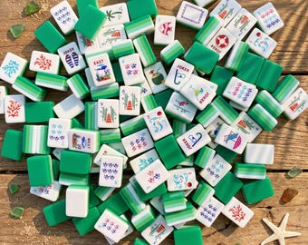 May include: A collection of green and white Mahjong tiles scattered on a wooden surface. The tiles feature various symbols, numbers, and characters, including "JOKER" and directional markers like "EAST" and "WEST". Some tiles have illustrations of fish, palm trees, and other designs.