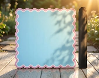 May include: A square light blue board with a scalloped edge featuring a pink and white border. Beside the board stands a tall, slender, black cylindrical case. The items are positioned on a wooden surface with natural outdoor lighting.