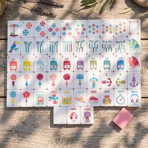 May include: A complete set of white Mahjong tiles with colorful illustrations. The tiles feature various symbols, including umbrellas, flowers, suitcases, and dragons. A pink eraser is also present on the wooden surface.