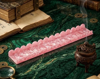 May include: A rectangular incense holder made of pink marbled acrylic with a scalloped edge design. The holder features a flat base and a raised back panel with a series of rounded, semi-circular cutouts along the top edge, creating a decorative border.